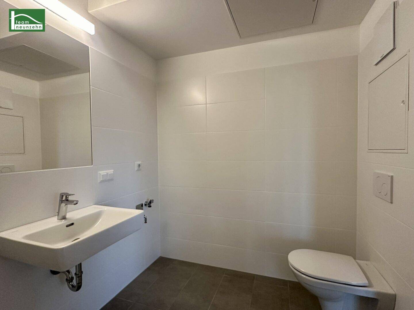 Helles Badezimmer mit WC, Waschbecken und großen weißen Wandfliesen, sehr sauber.