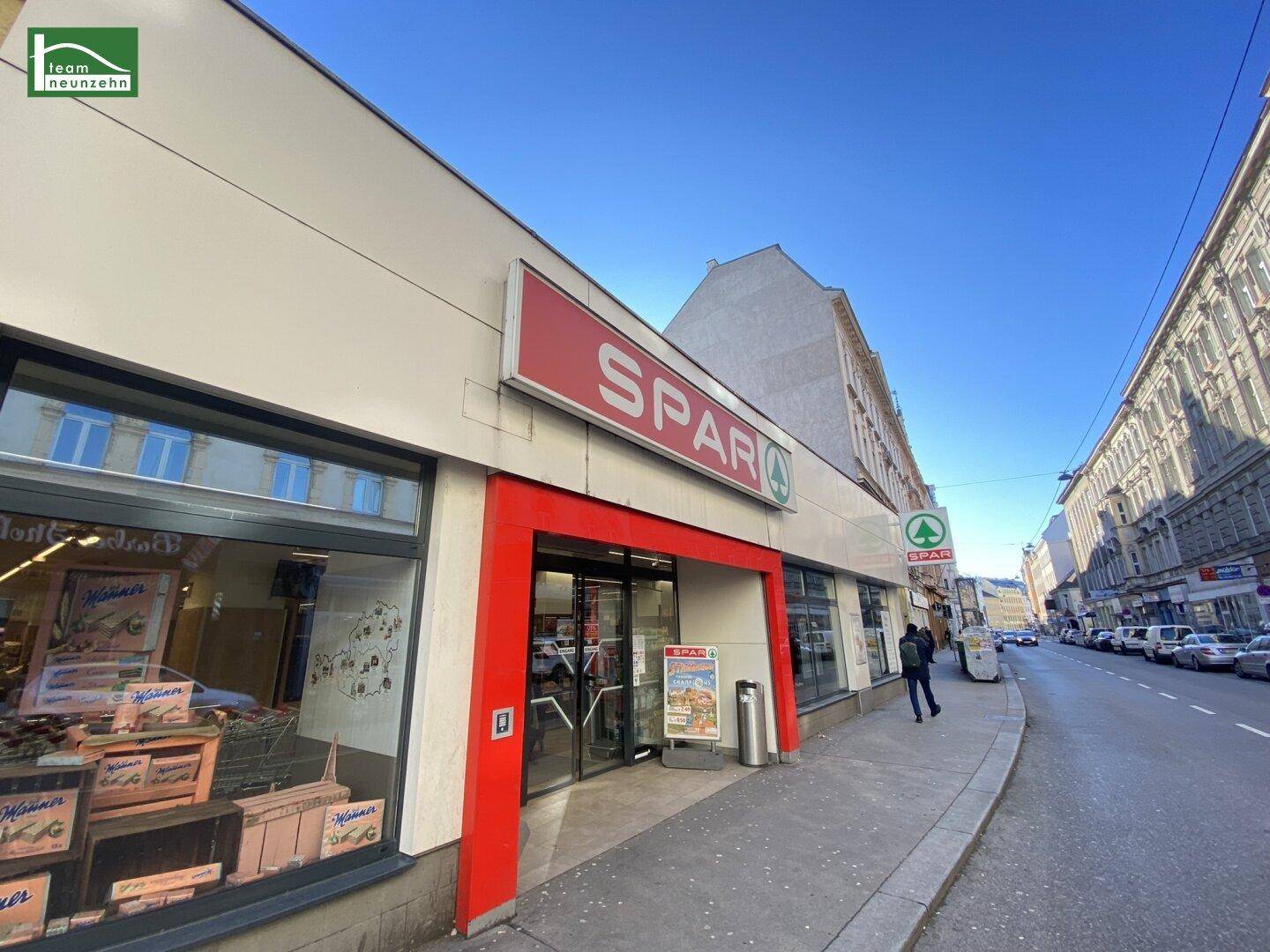 Eingang eines SPAR-Supermarktes an einer belebten Stadtstraße mit weiteren Gebäuden im Hintergrund.