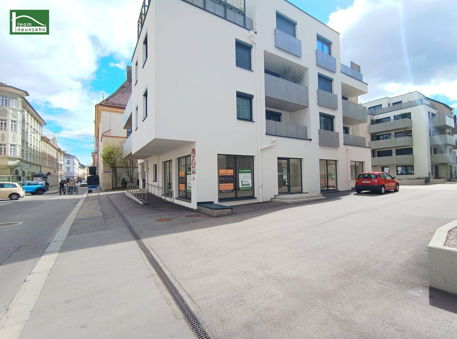 Modernes Gebäude mit weißer Fassade, Balkonen und Geschäftsflächen im Erdgeschoss an einer Straße.