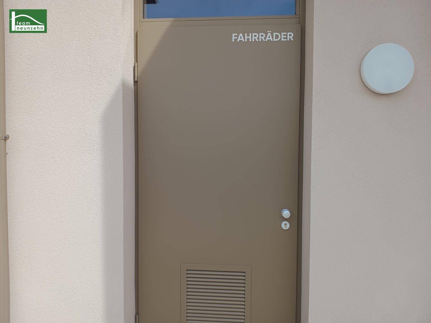 Eingang zum Fahrradraum mit brauner Tür und Lüftungsgitter, außen am Gebäude gelegen.