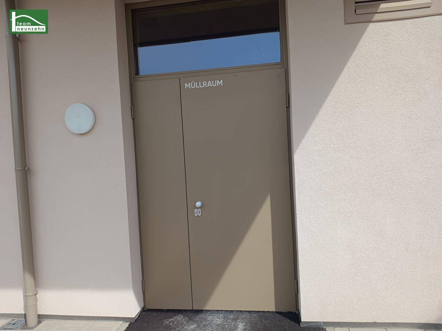 Eingang zum Müllraum mit brauner Tür und kleinem Fenster, außen am Gebäude gelegen.