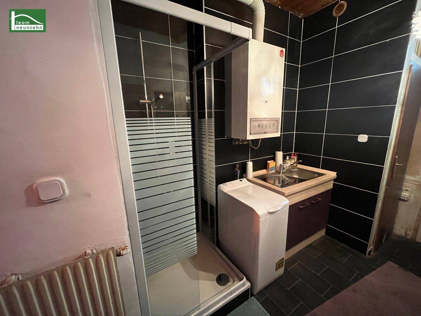 Badezimmer mit schwarzem Fliesenboden, Duschkabine, Waschbecken und Warmwasserbereiter, integriert in den Raum.