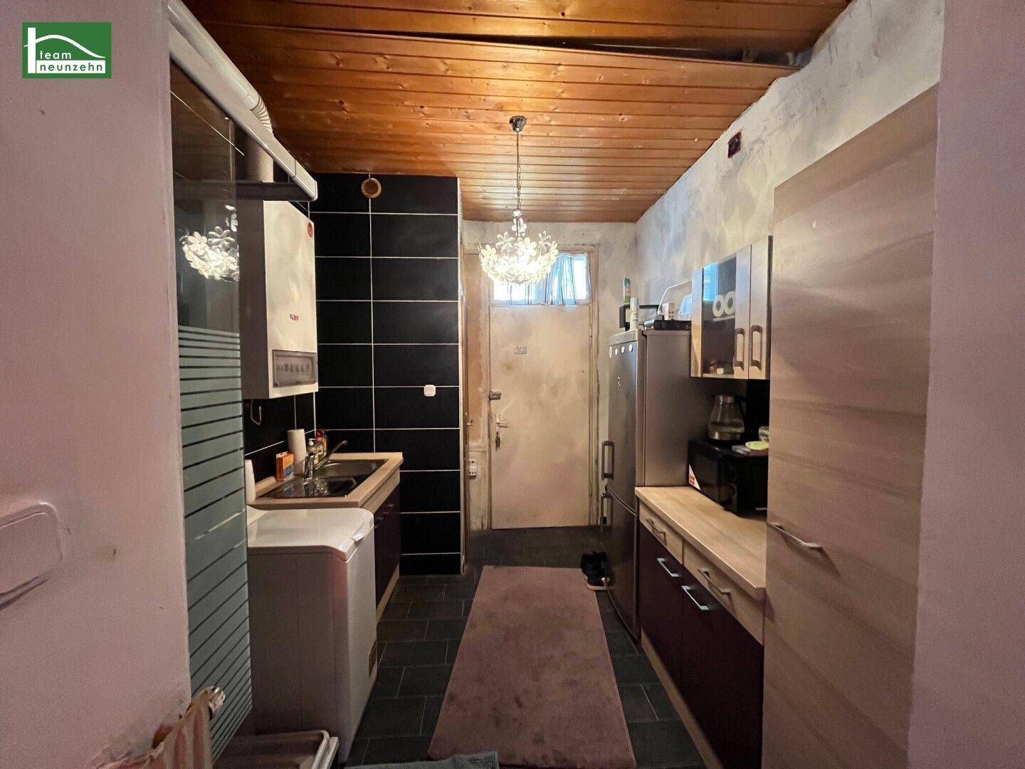 Schmale Küche mit schwarzen Fliesen, Waschmaschine, Kühlschrank und Holzdecke, die eine funktionale Kochzone bietet.