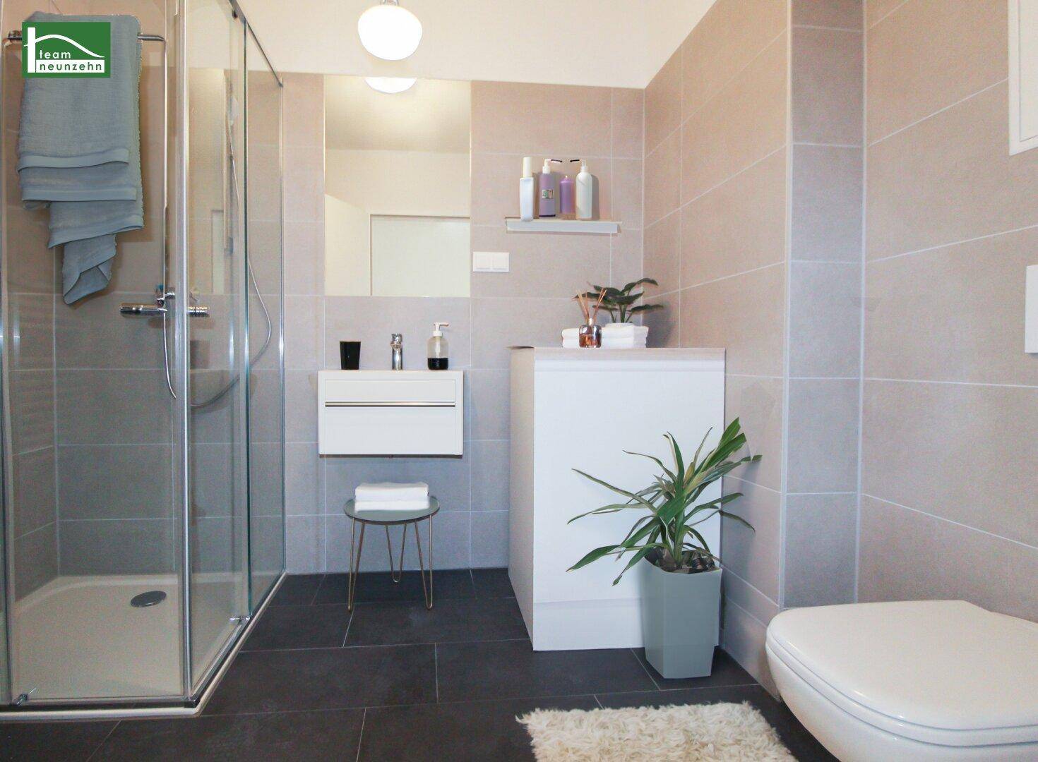 Modernes Badezimmer mit grauen Fliesen, Duschkabine, wandhängendem Waschbecken und Toilette.
