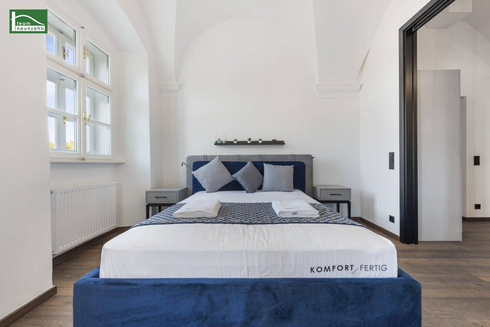 Helles Schlafzimmer mit blauem Bett, Holzboden und hohen Gewölbedecken.