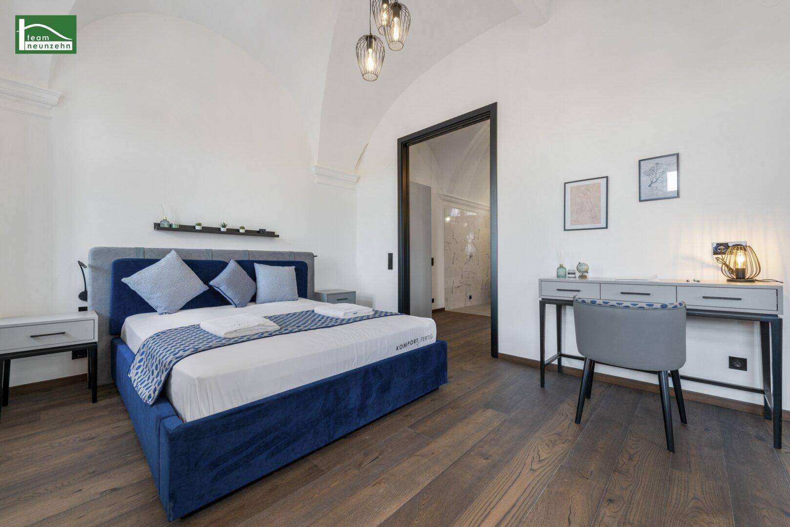 Schlafzimmer mit blauem Bett, Schreibtisch und Holzboden, verbunden mit einem weiteren Raum.
