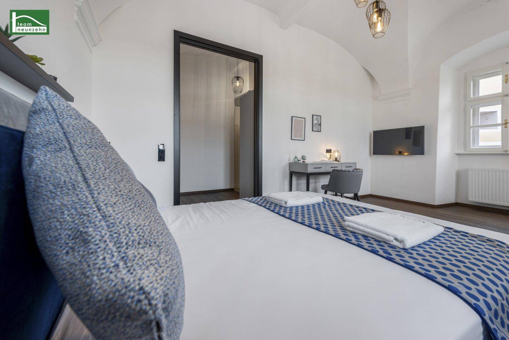 Gemütliches Schlafzimmer mit blauem Bett, Holzboden und Blick auf den Fernseher.