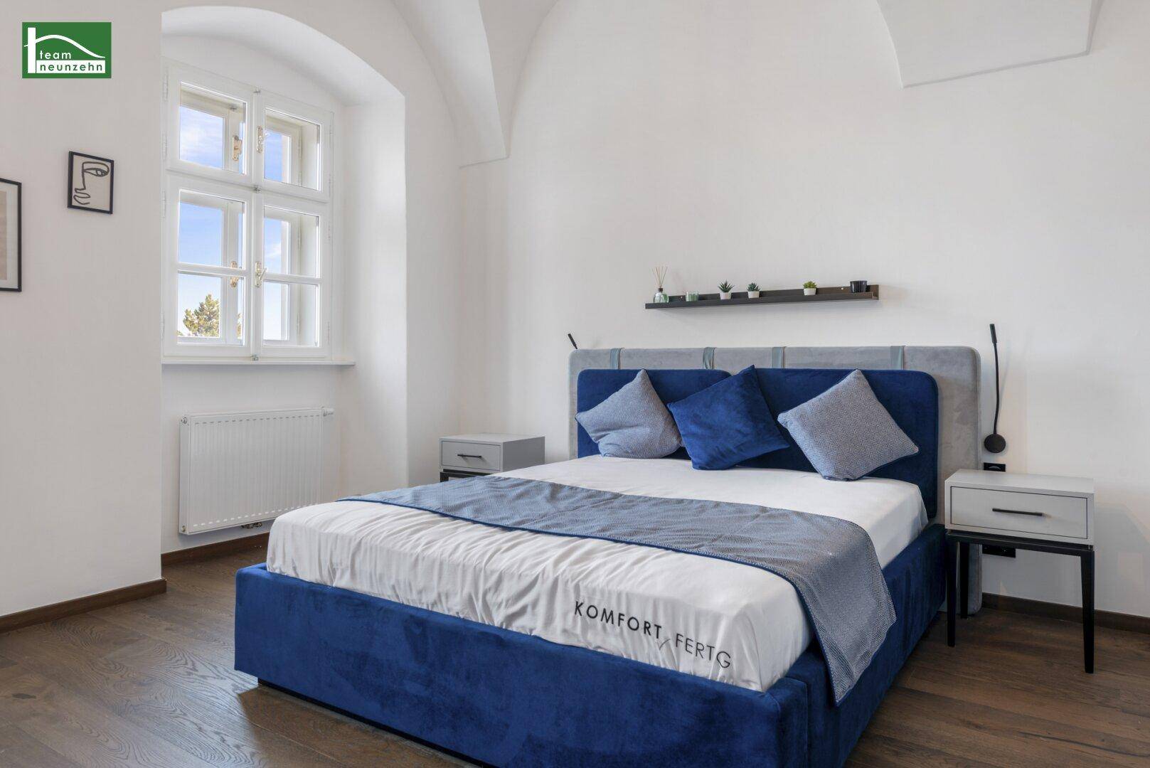 Helles Schlafzimmer mit blauem Bett, Holzboden und großen Fenstern.