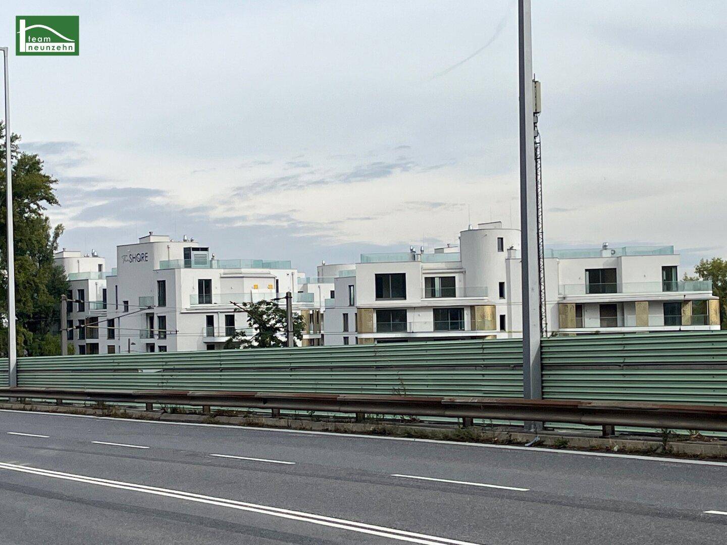 Moderne weiße Gebäude mit großen Fenstern und Flachdächern, hinter einer grünen Lärmschutzwand.