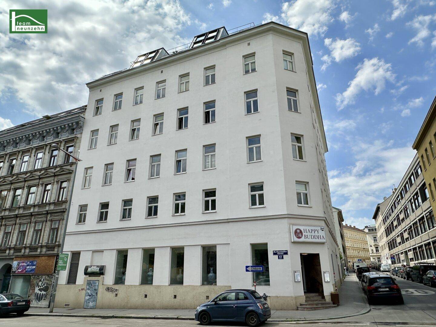 Weißes Eckgebäude in einer städtischen Umgebung mit vielen Fenstern und Geschäften im Erdgeschoss.