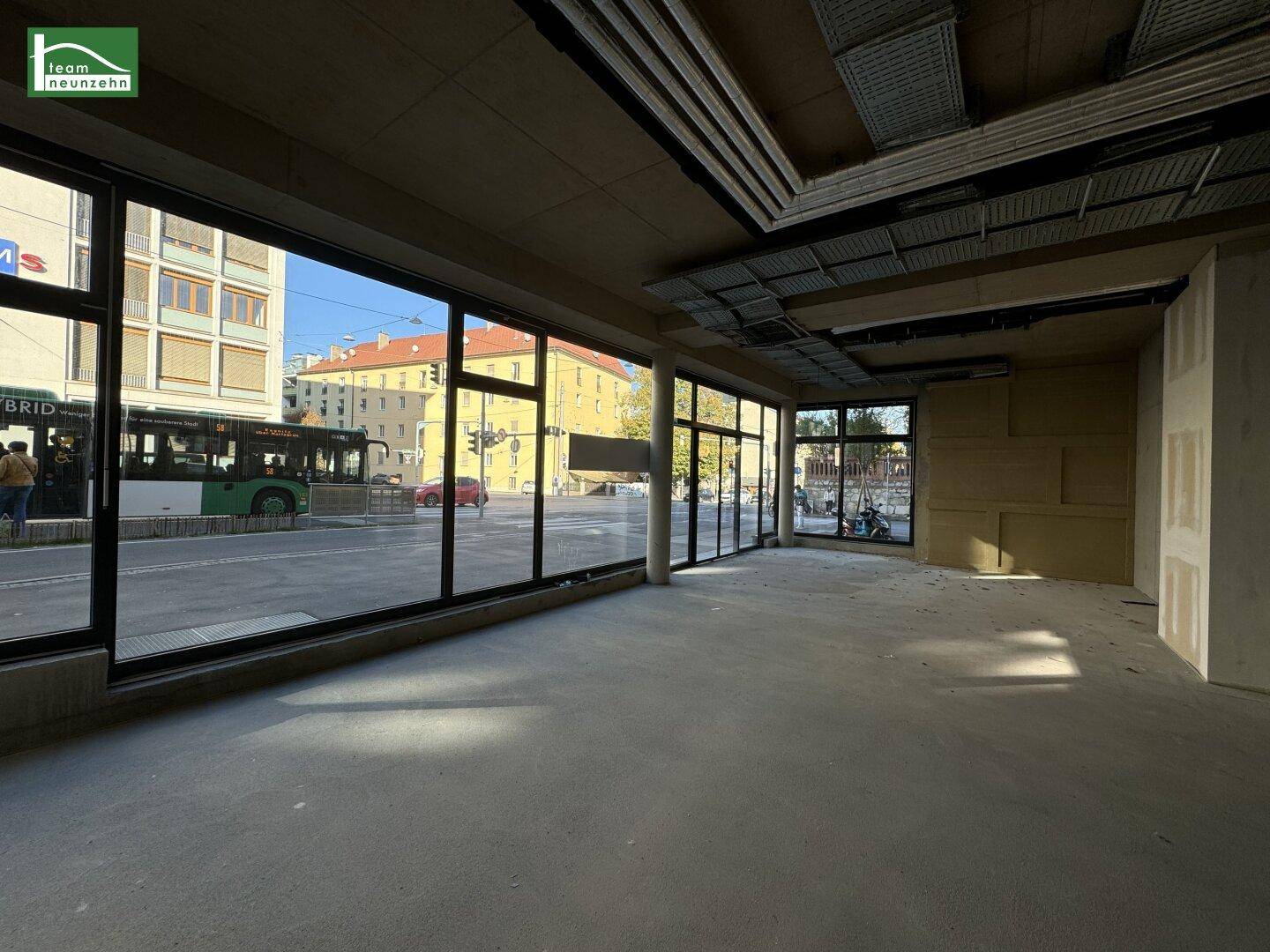 Unfertiger Gewerberaum mit großen Schaufenstern zur Straße, viel Tageslicht und Betonboden.