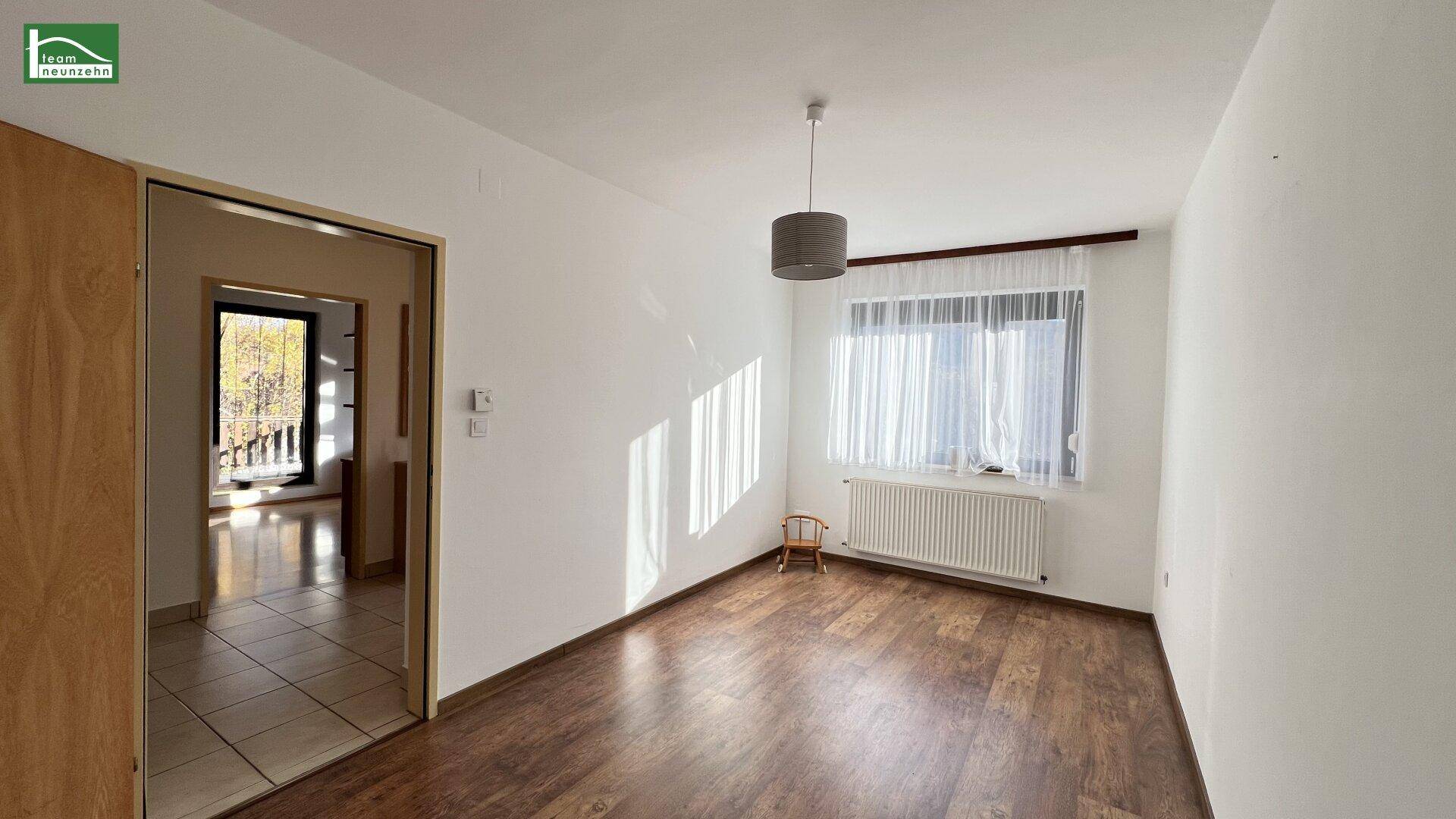 Helles Wohnzimmer mit dunklem Laminatboden und großem Fenster, das viel Licht hereinlässt.