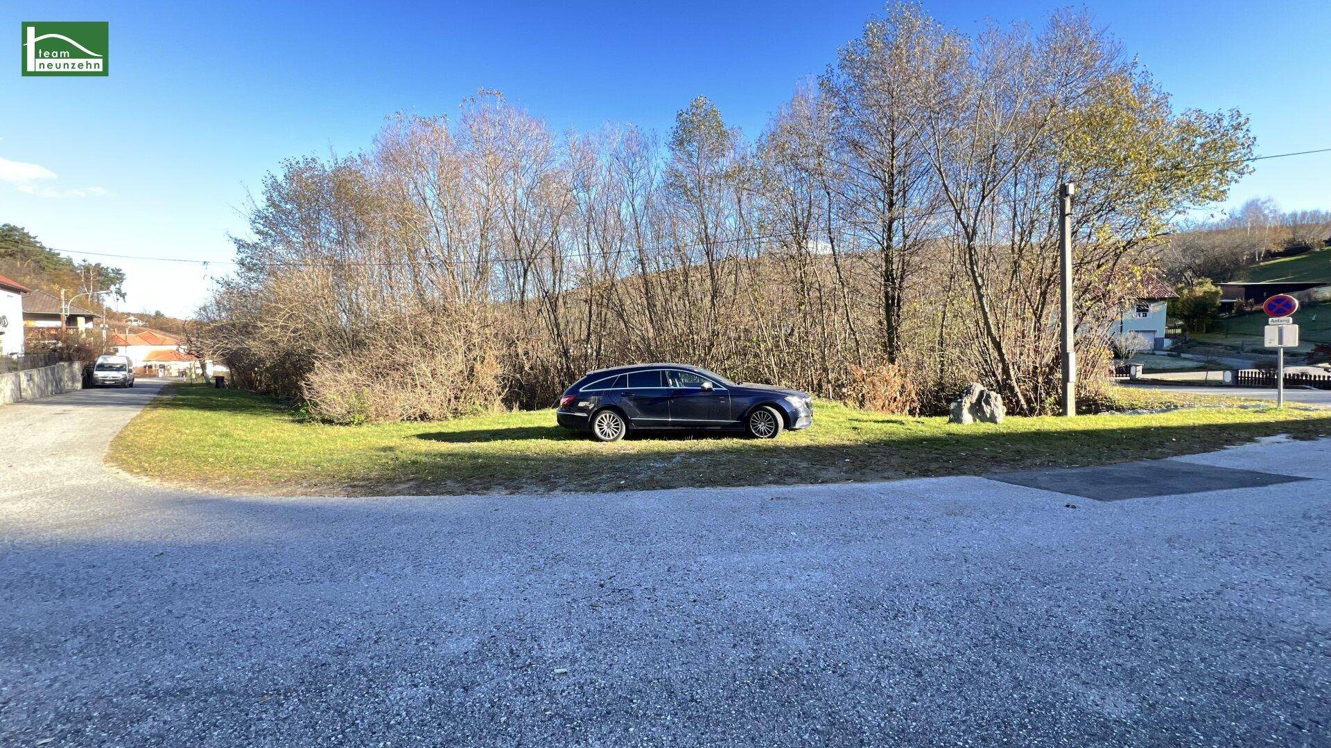 Blick auf ein Grundstück mit einem geparkten Auto, umgeben von Bäumen und Sträuchern unter blauem Himmel.