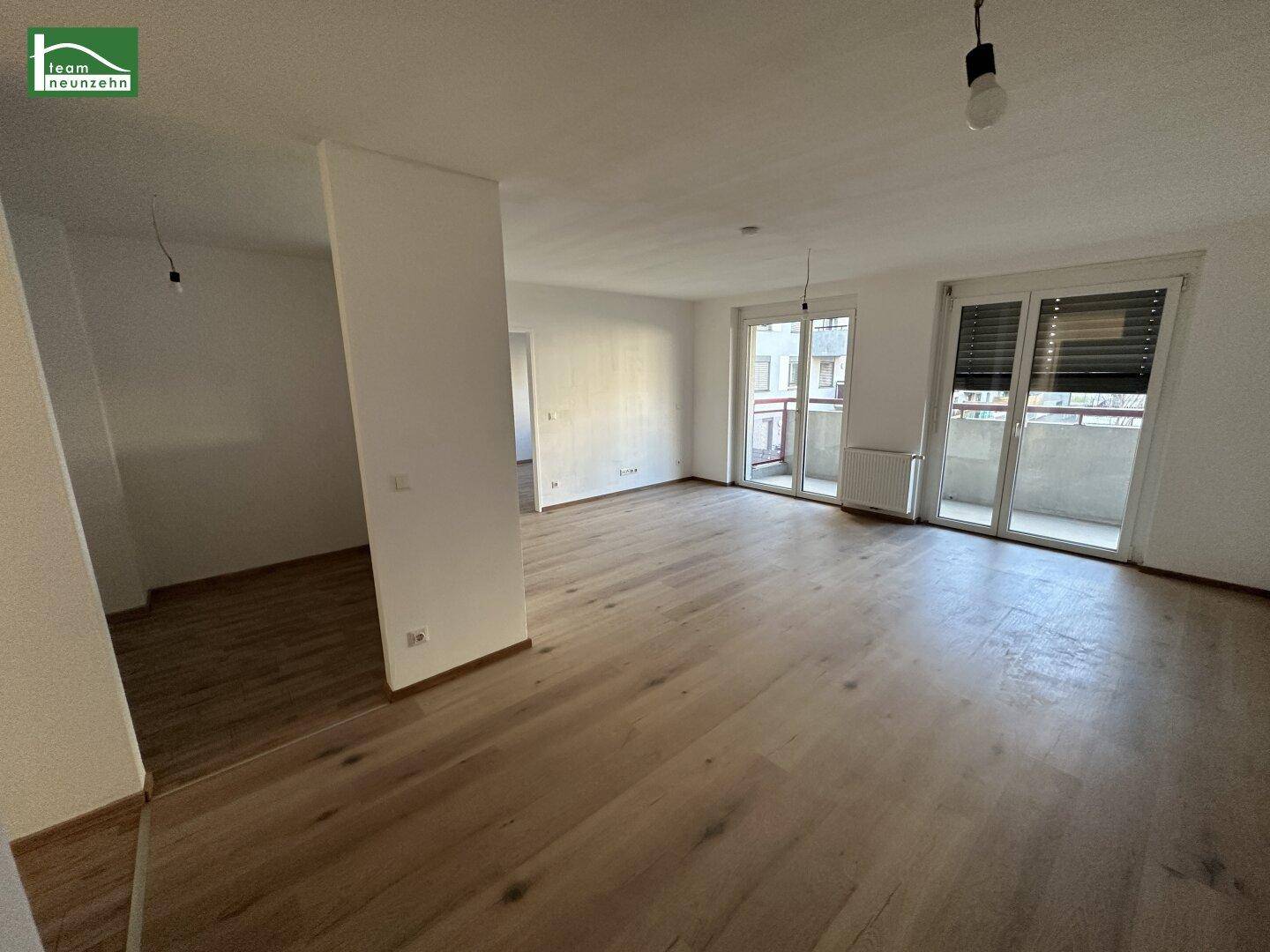 Geräumiges Wohnzimmer mit Holzboden, großen Fenstern und Zugang zu einem Balkon.