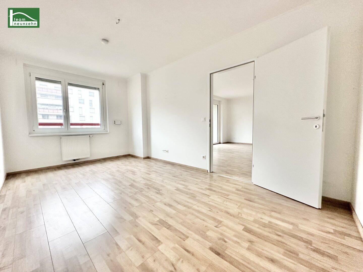 Helles Zimmer mit Fenster und Holzboden, durch eine offene Tür mit einem weiteren Raum verbunden.