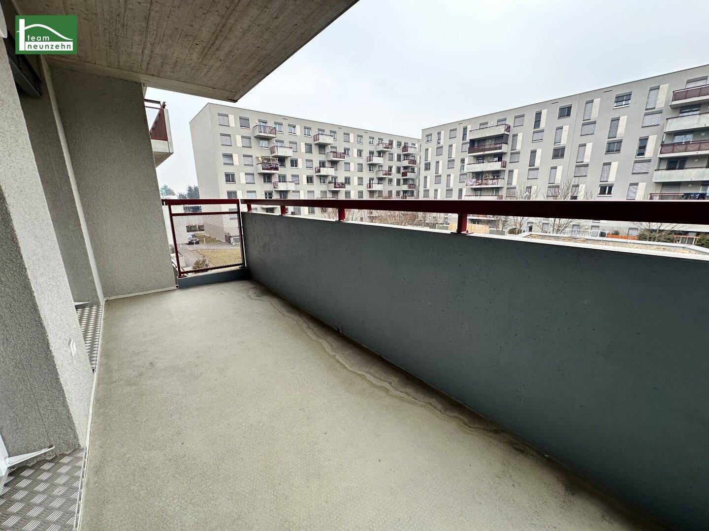 Betonbalkon mit rotem Geländer und Blick auf umliegende Wohngebäude in städtischer Umgebung.