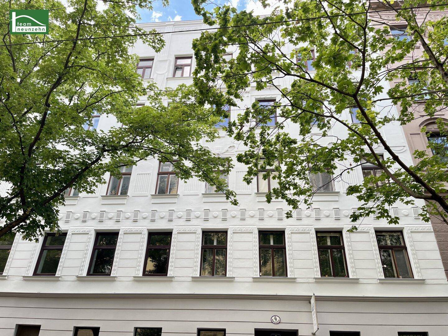 Die weiße, reich verzierte Fassade eines Altbaus mit großen Fenstern und grünen Bäumen davor.