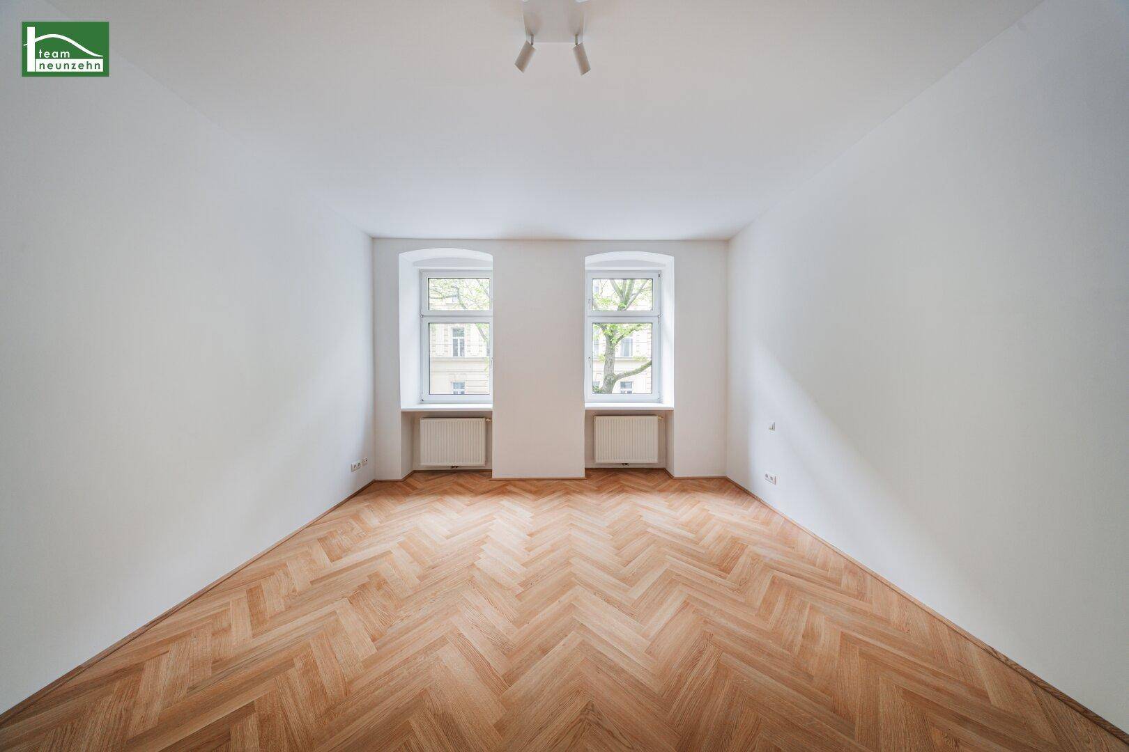 Heller, leerer Raum mit Fischgrätparkettboden und zwei Fenstern, die viel Tageslicht hereinlassen.