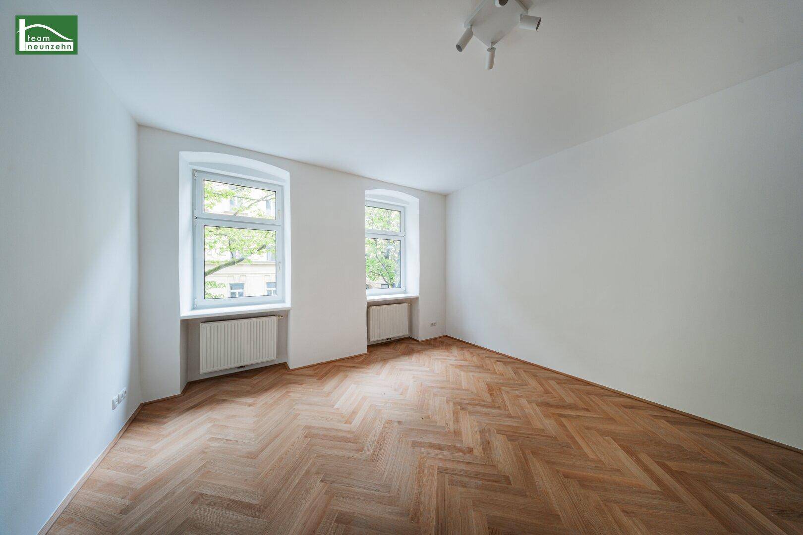 Geräumiges Zimmer mit klassischem Fischgrätparkett und zwei großen Fenstern mit Blick ins Grüne.