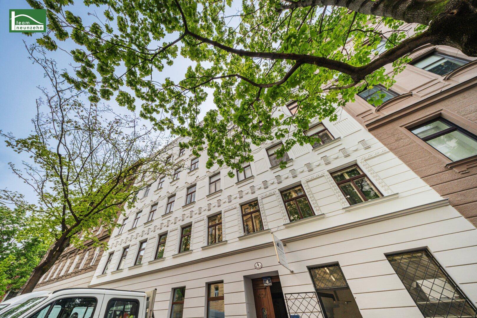 Ansicht der weißen Gebäudefassade mit traditionellen Fenstern und Bäumen, die das obere Stockwerk umrahmen.