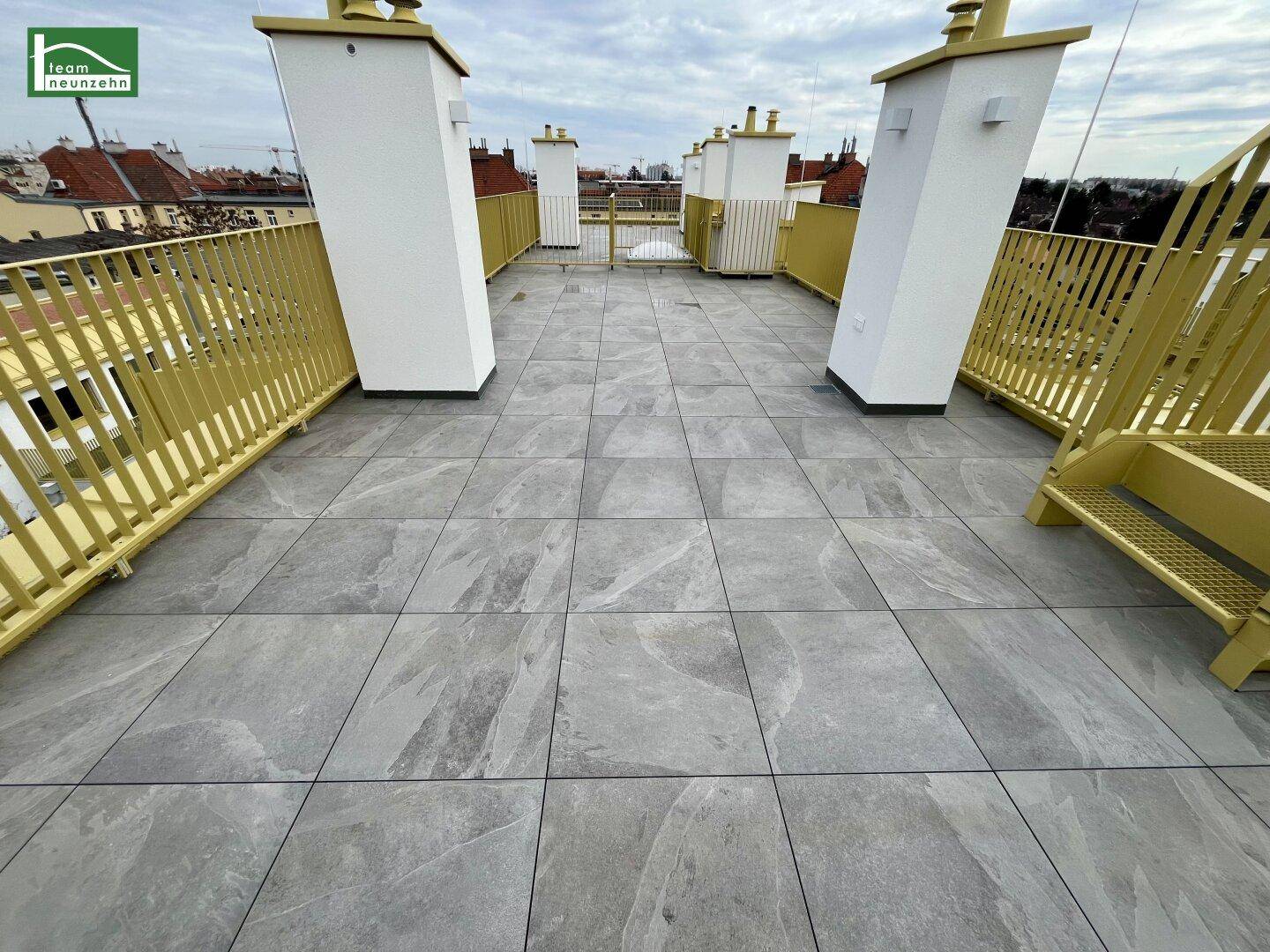Geräumige Dachterrasse mit grauen Fliesen und gelbem Geländer, bietet Stadtblick.