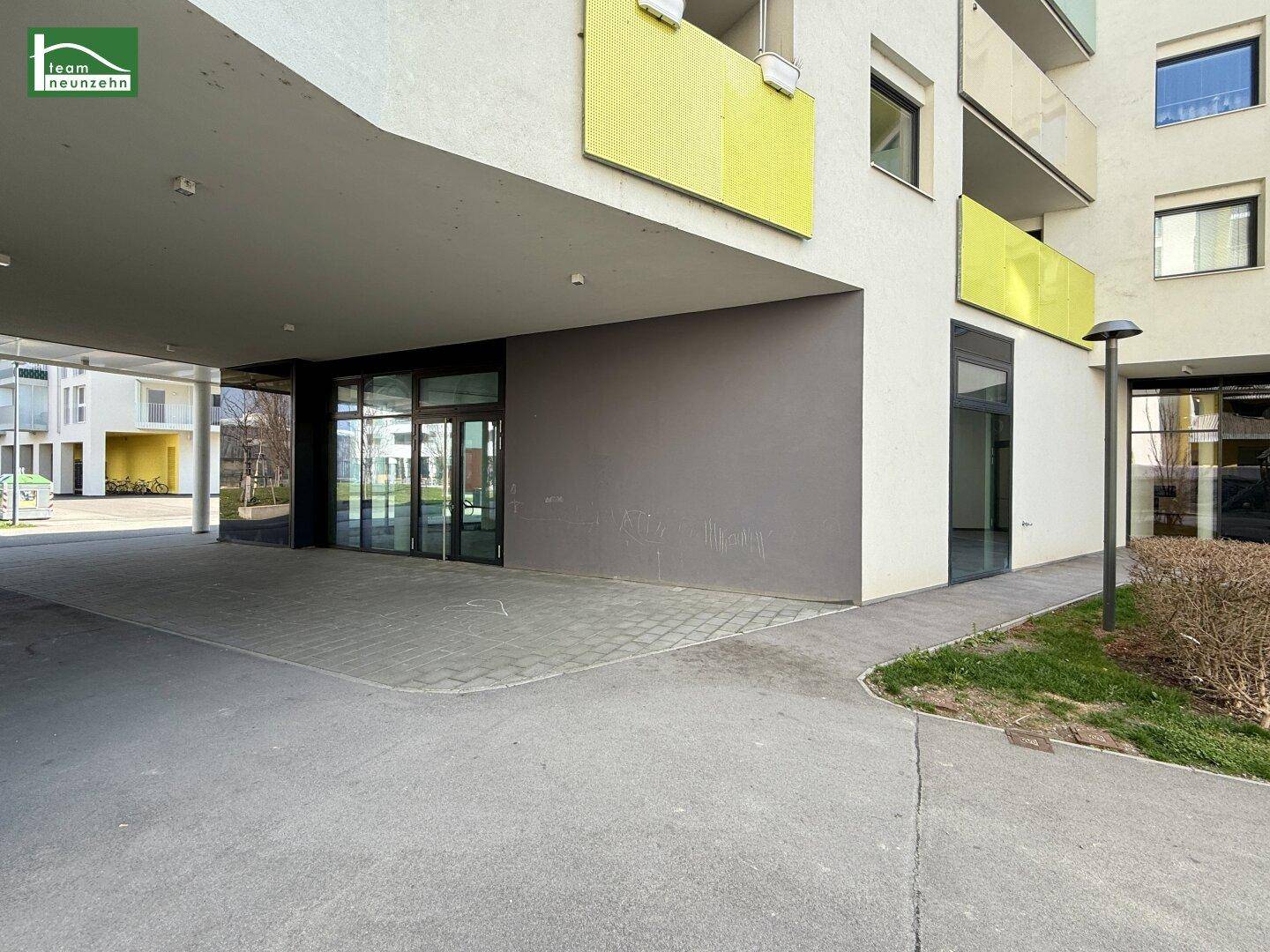 Überdachter Eingangsbereich eines modernen Wohn- und Geschäftsgebäudes mit gepflastertem Gehweg.
