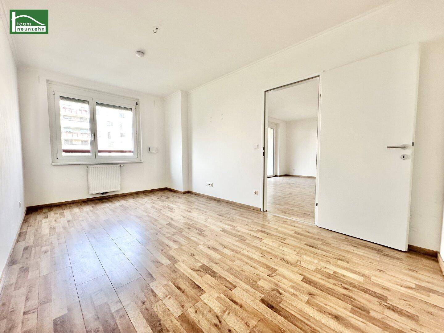 Helles Zimmer mit Fenster, Heizkörper und Laminatboden, bietet Zugang zu einem weiteren Raum.