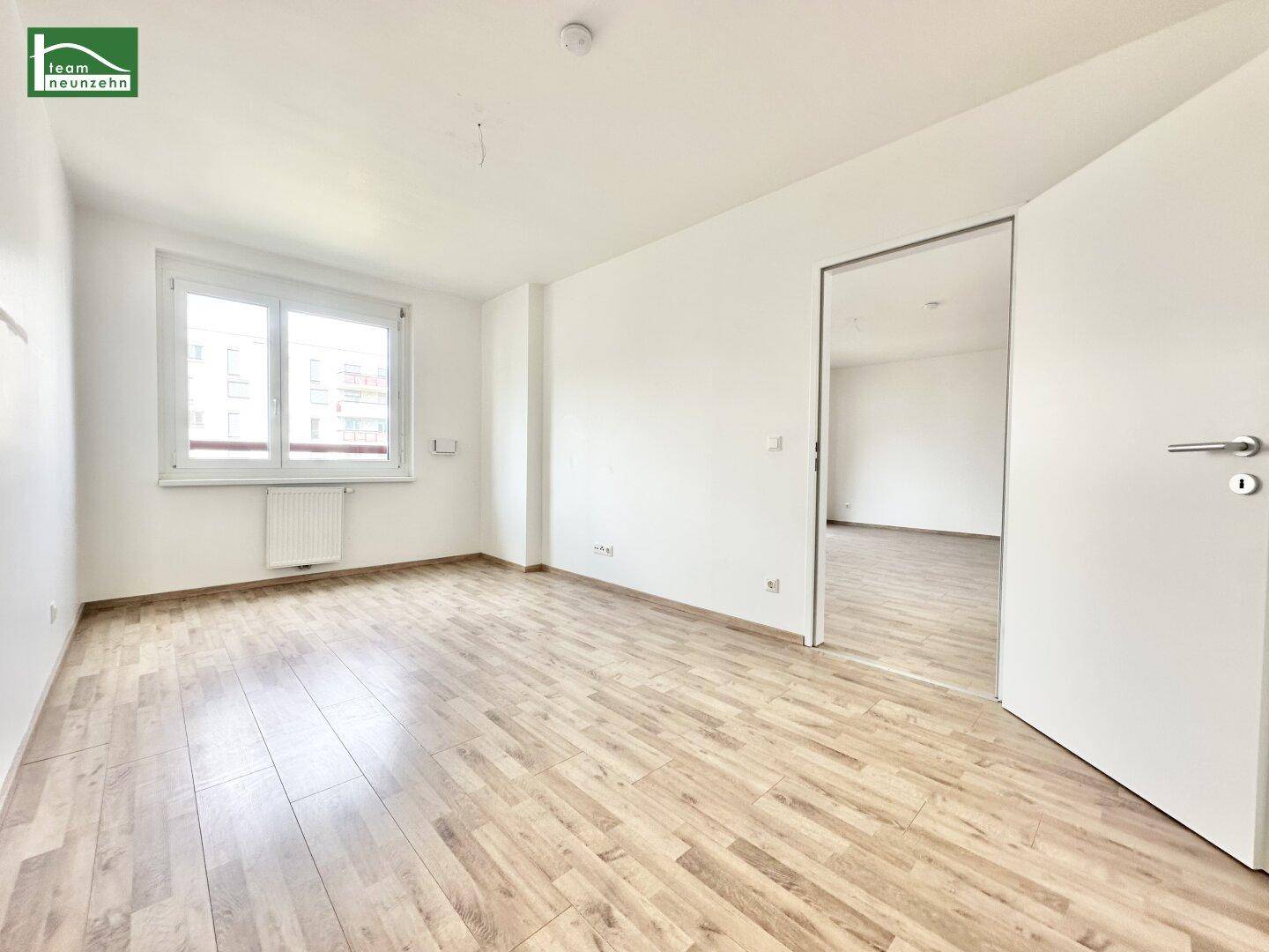 Helles, leeres Zimmer mit Laminatboden und einem großen Fenster, das viel Tageslicht hereinlässt.