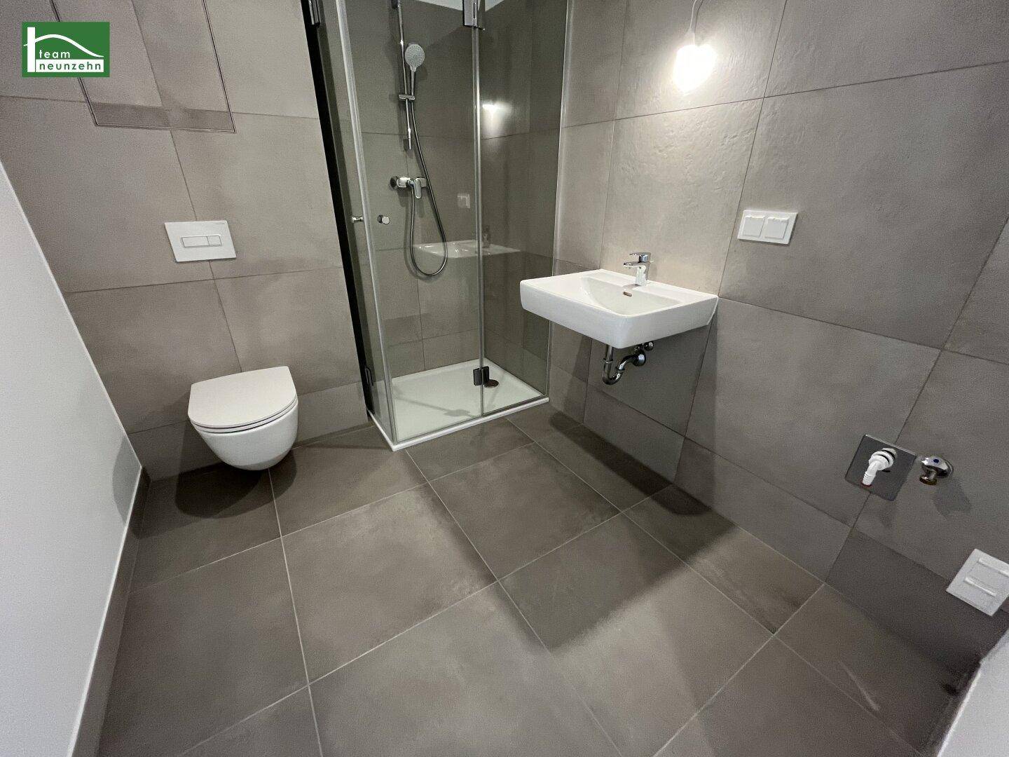 Modernes Badezimmer mit grauen Fliesen, wandhängendem WC und begehbarer Dusche.
