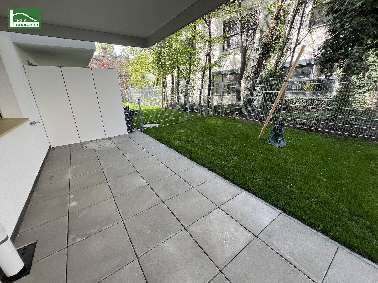 Außenbereich mit großer gefliester Terrasse und grünem Rasen, umgeben von Bäumen.