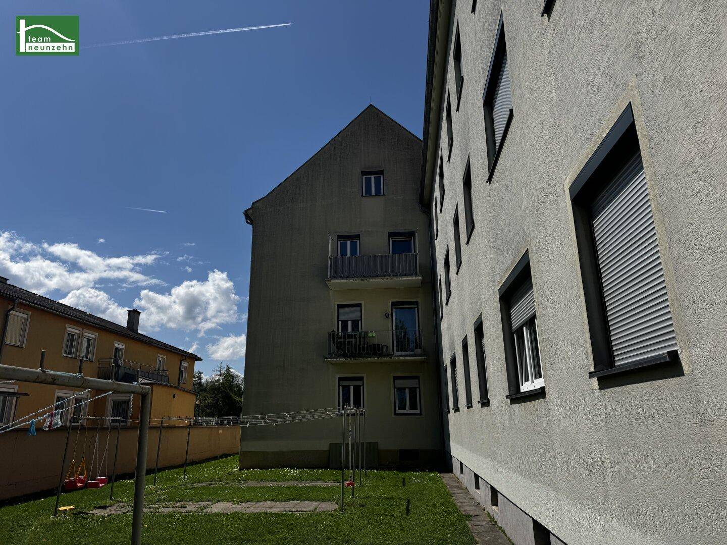 Mehrfamilienhaus mit hellgrüner Fassade und einem kleinen Spielplatz im Vorgarten bei Sonnenschein.