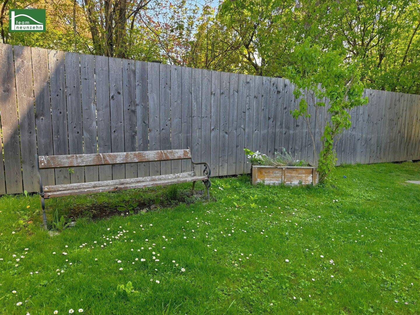 Grüner Gartenbereich mit einer Holzbank, einem Holzzaun und einem kleinen Pflanzenkübel.
