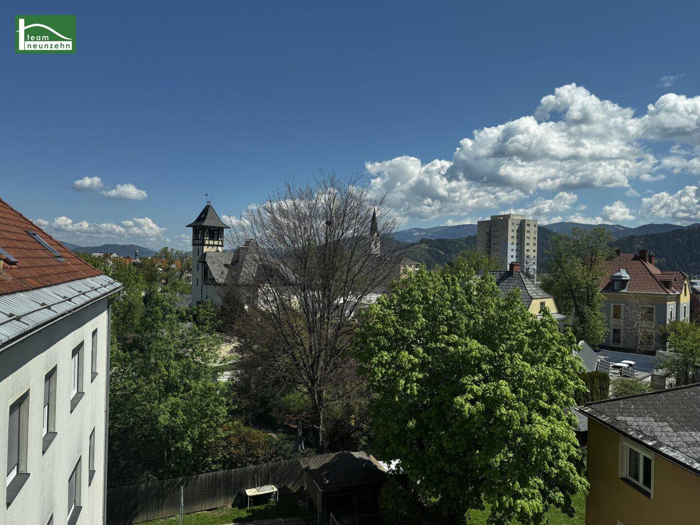 Panoramablick über eine Stadt mit historischen Gebäuden, viel Grün und Bergen im Hintergrund.