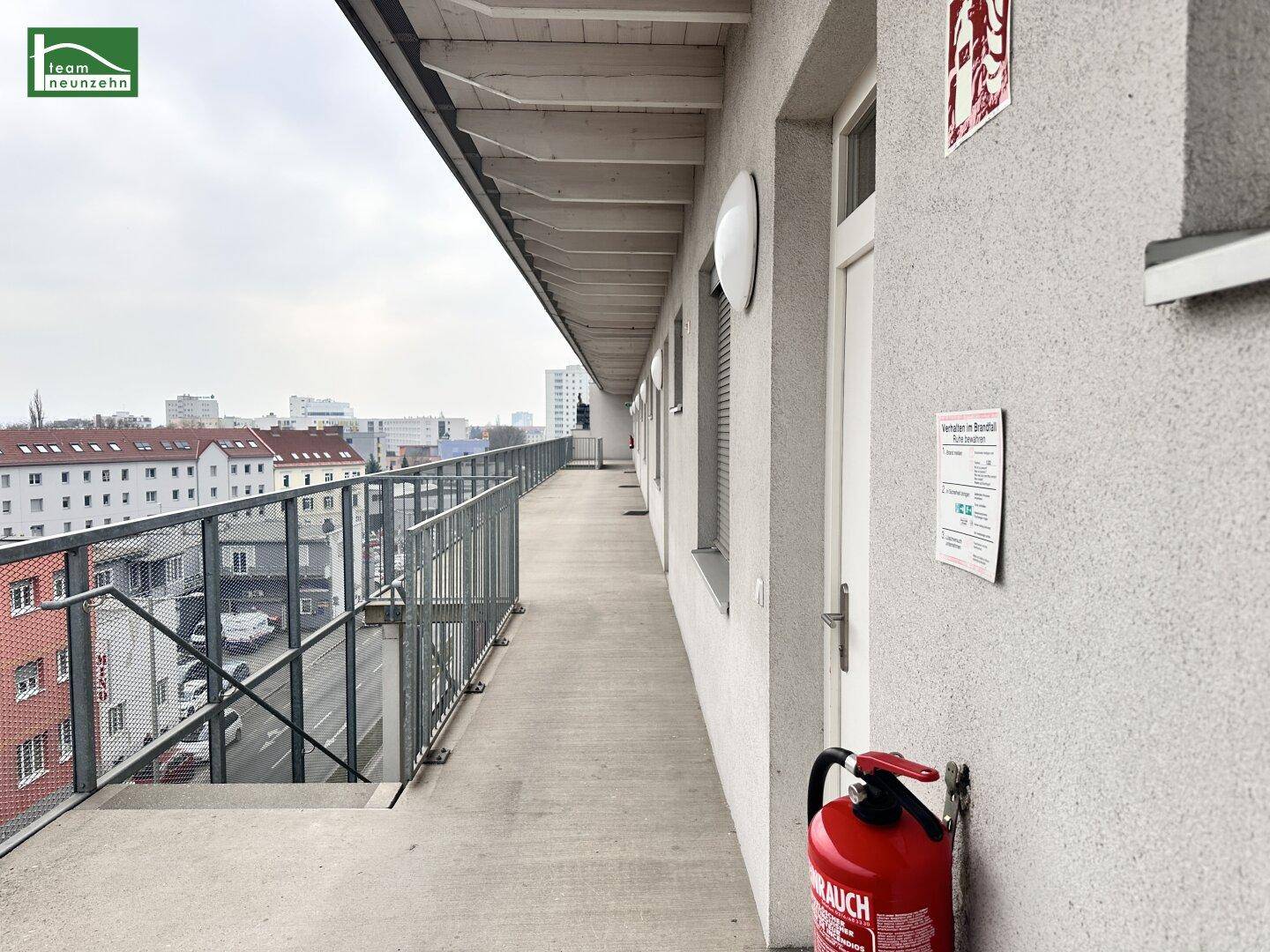 Langer Außenflur mit Metallgeländer, Feuerlöscher und Blick auf die umliegende Stadtbebauung.