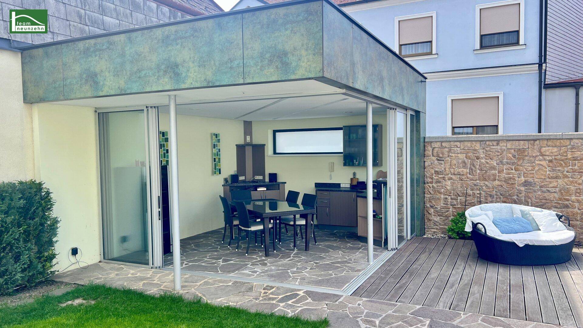 Überdachter Außenbereich mit Esstisch, Holzterrasse und angrenzendem Rasen.