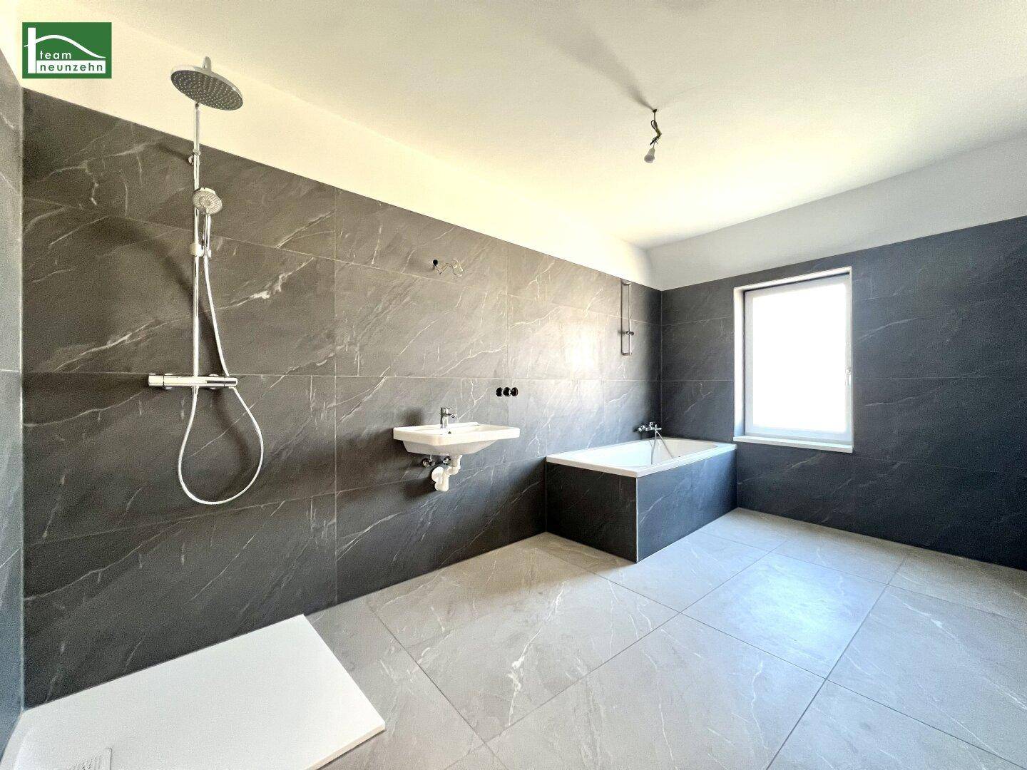 Geräumiges Badezimmer mit modernen grauen Fliesen, Badewanne, Regendusche und Fenster.