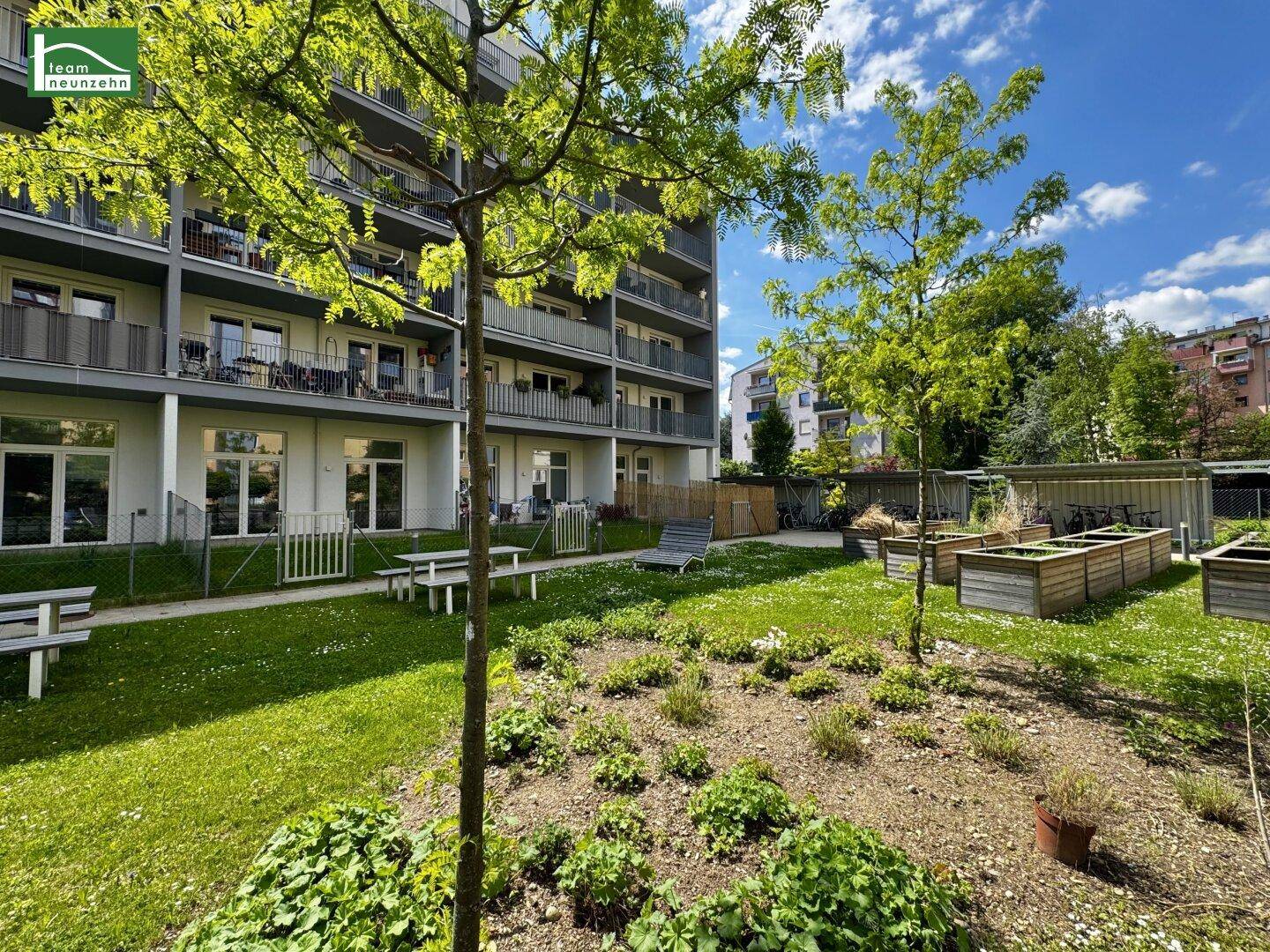Gemeinschaftlicher Gartenbereich mit Sitzbänken und jungen Bäumen vor dem modernen Apartmentgebäude.