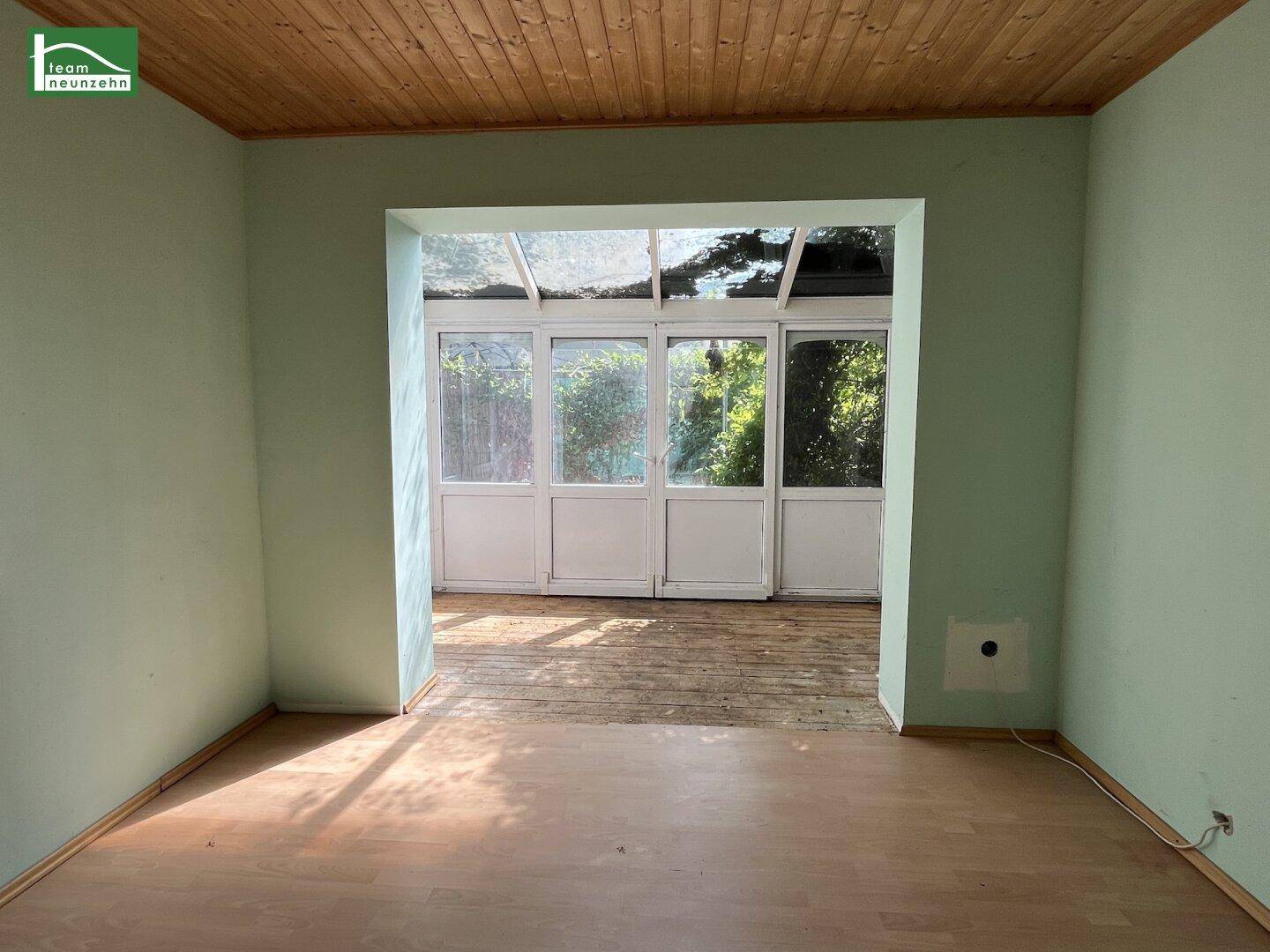 Wohnraum mit Holzdecke und Laminatboden, der in einen hellen Wintergarten übergeht.