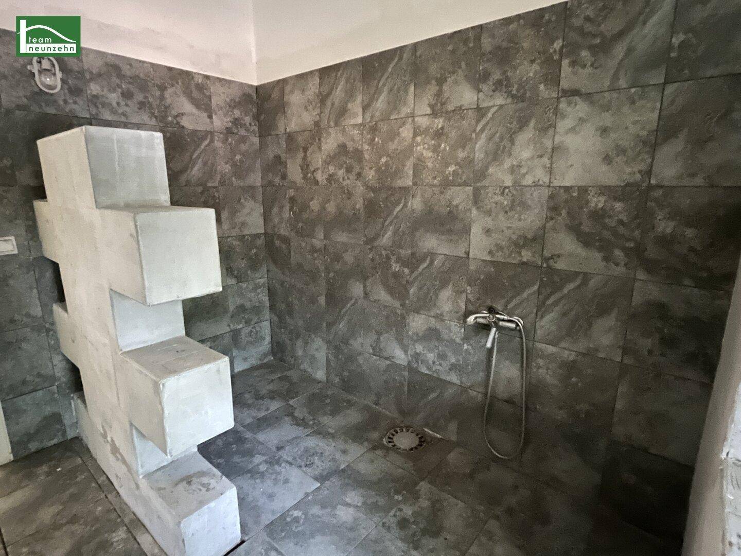 Badezimmer mit grauen Fliesen, Duschbereich und einer Duscharmatur an der Wand.
