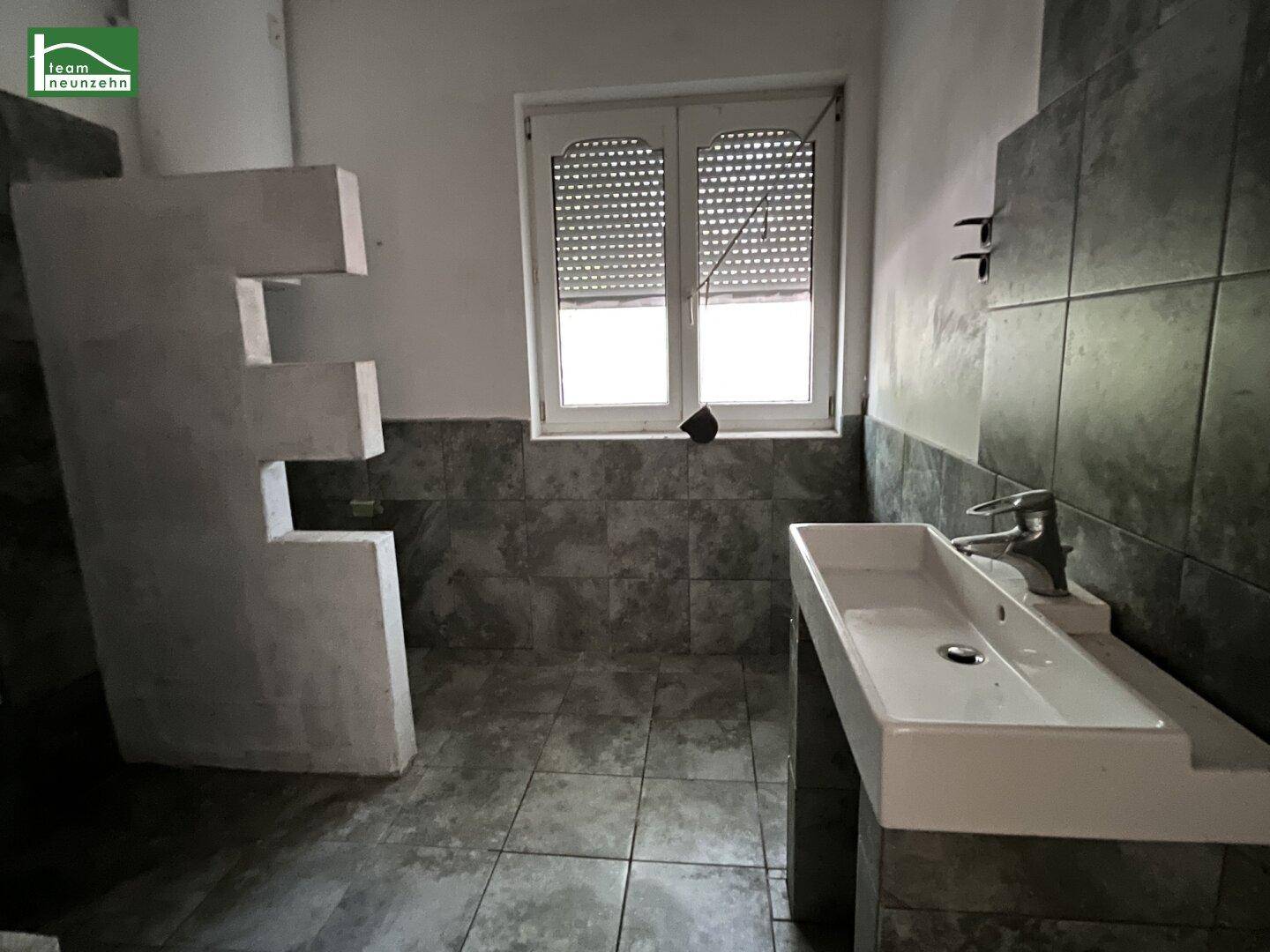 Badezimmer mit modernem Waschbecken, grauen Fliesen und einem Fenster mit Rollladen.
