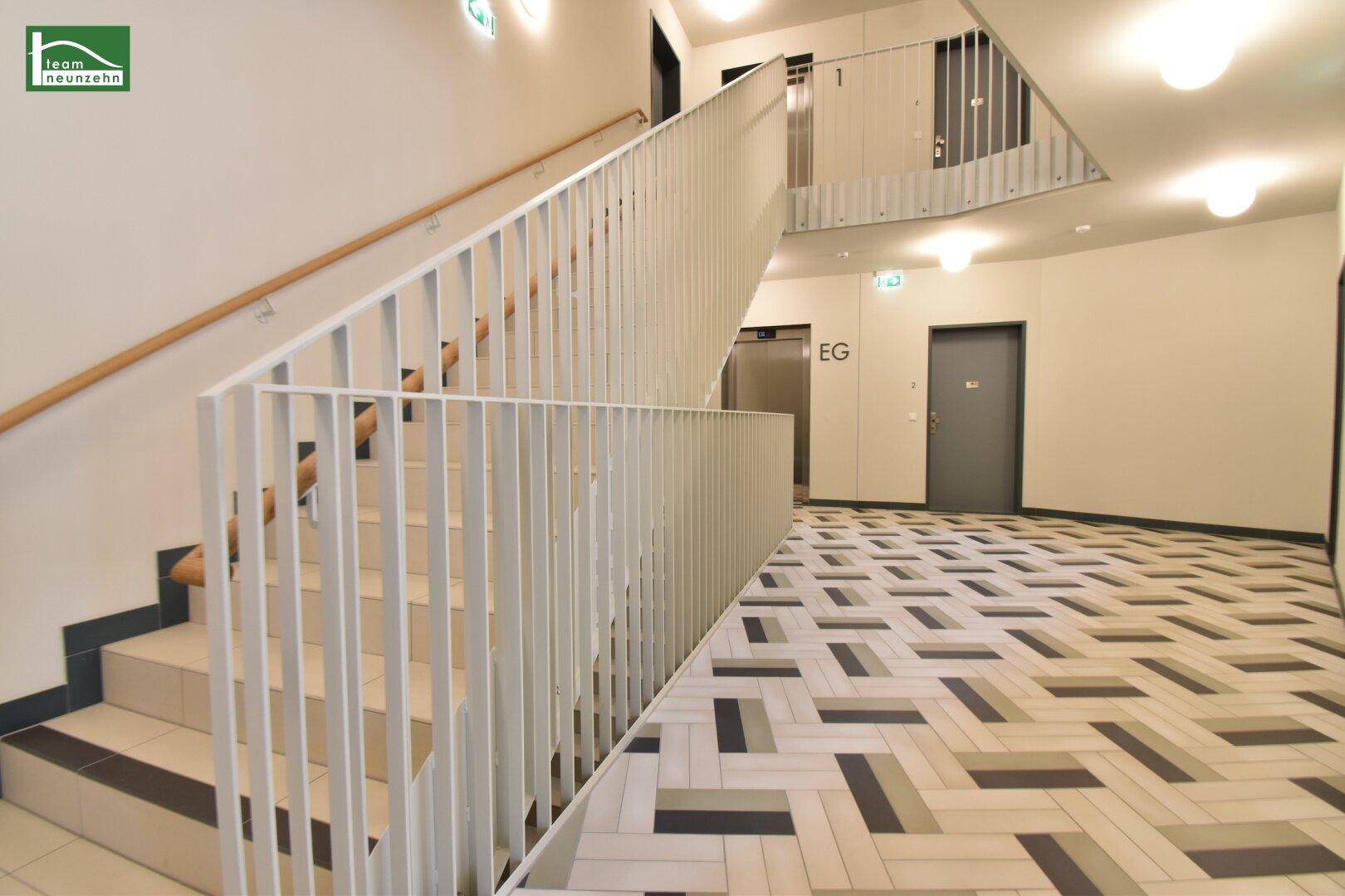 Modernes Treppenhaus mit hellem Geländer, Fliesenboden und Zugang zum Aufzug im Erdgeschoss.