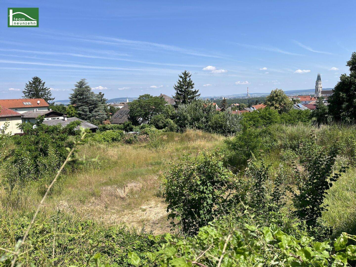 Panoramablick über eine grüne Wiese und Büsche mit Häusern und einem Kirchturm in der Ferne.