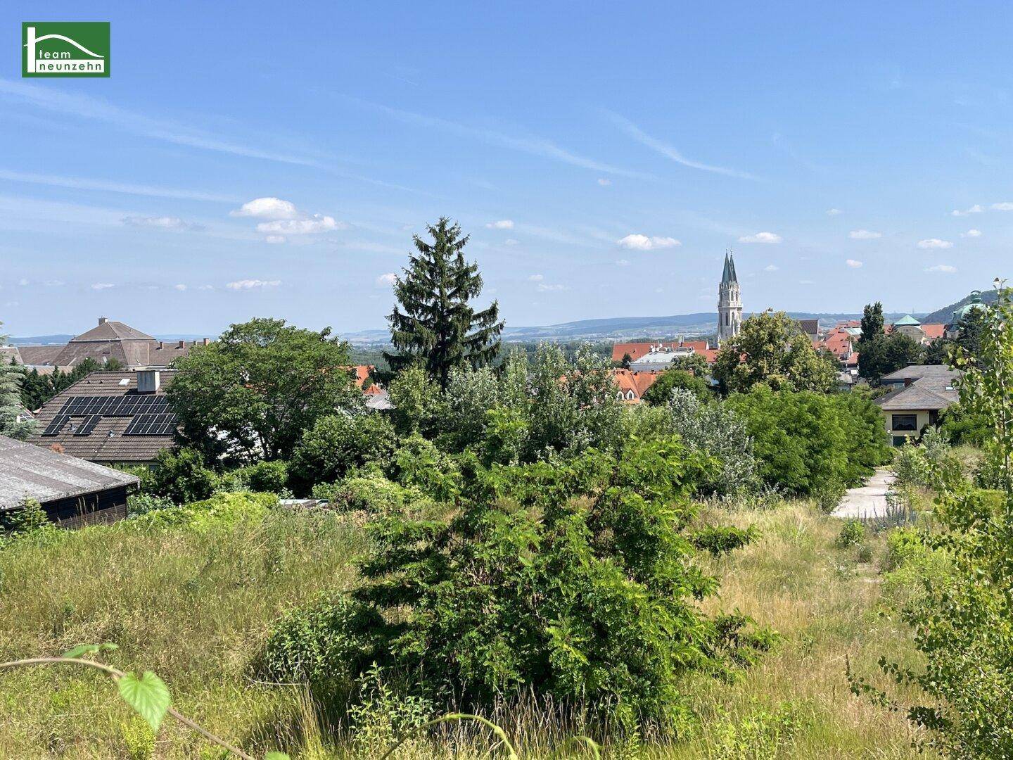 Ausblick auf eine hügelige Landschaft mit üppiger Vegetation, Dächern und einem Kirchturm am Horizont.