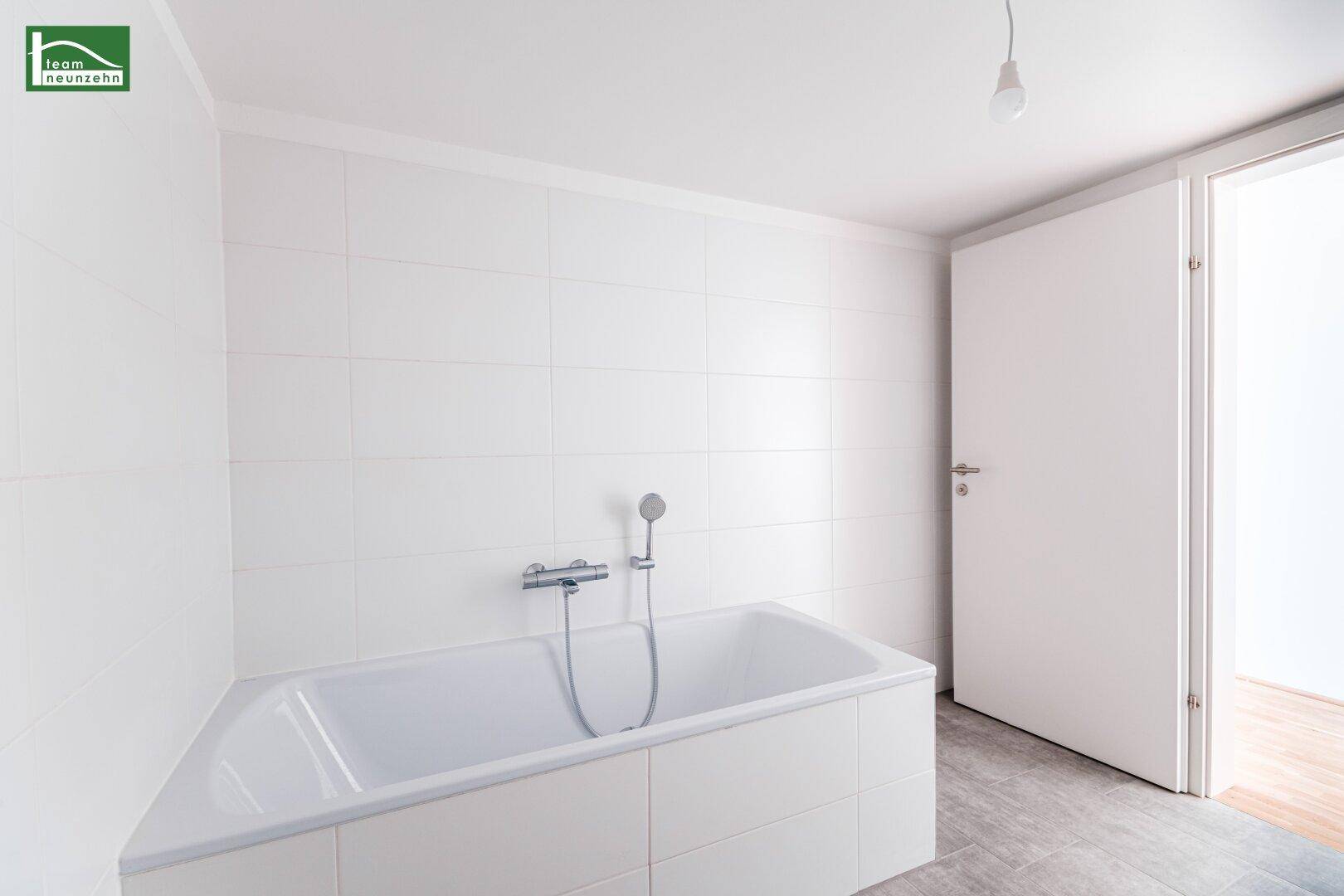 Helles Badezimmer mit Badewanne und weißen Fliesen an den Wänden und auf dem Boden.