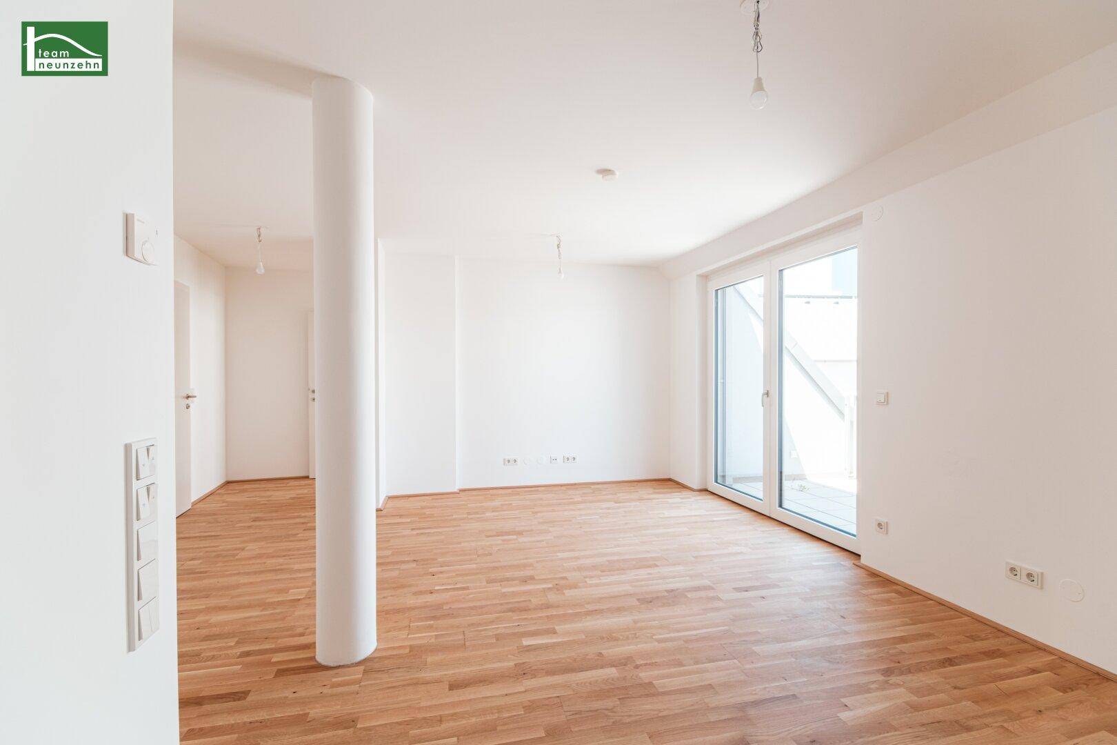 Heller, leerer Wohnraum mit Parkettboden und großem Fenster zum Außenbereich.