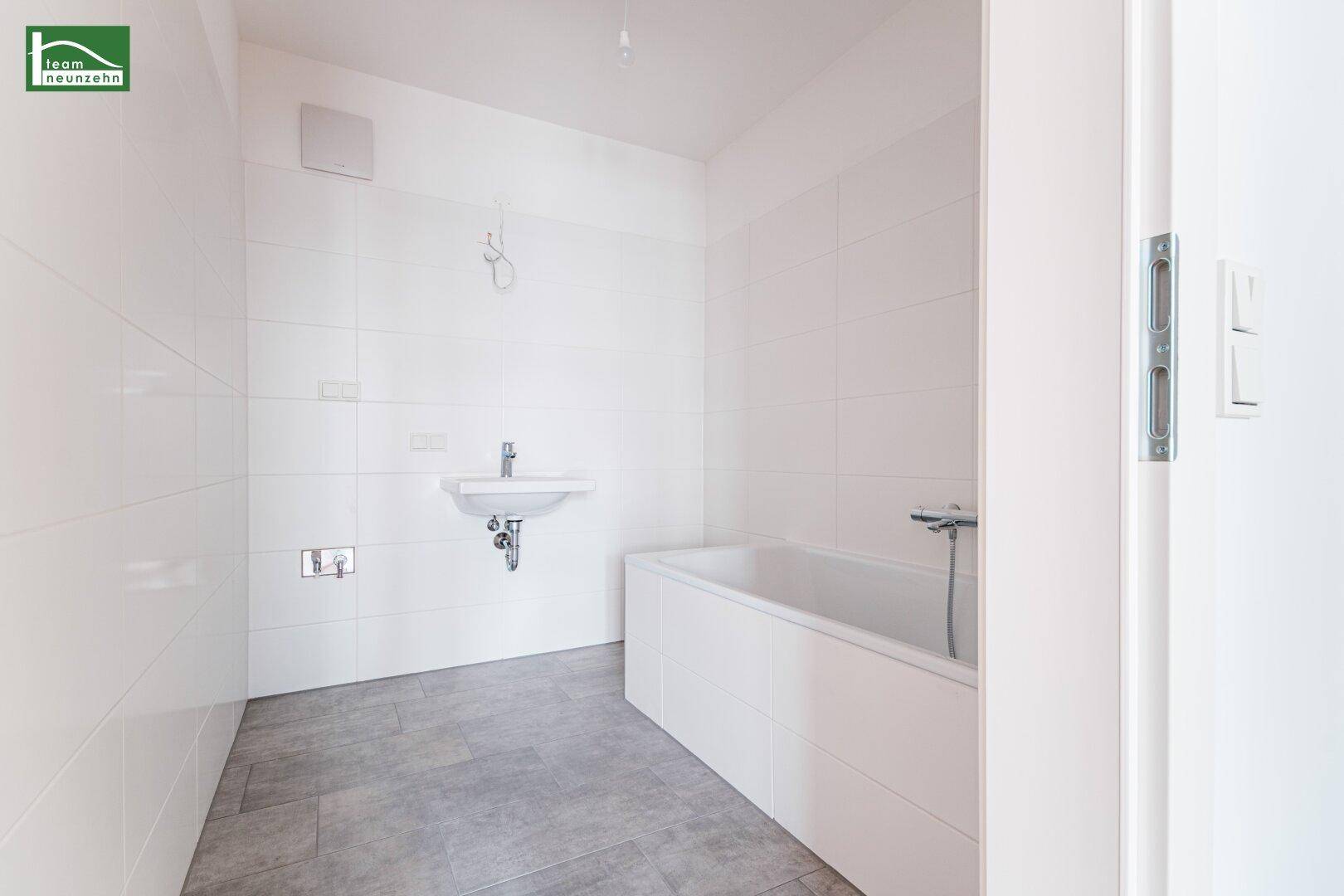 Helles Badezimmer mit Badewanne, Waschbecken, weißen Fliesen und Lüftung.
