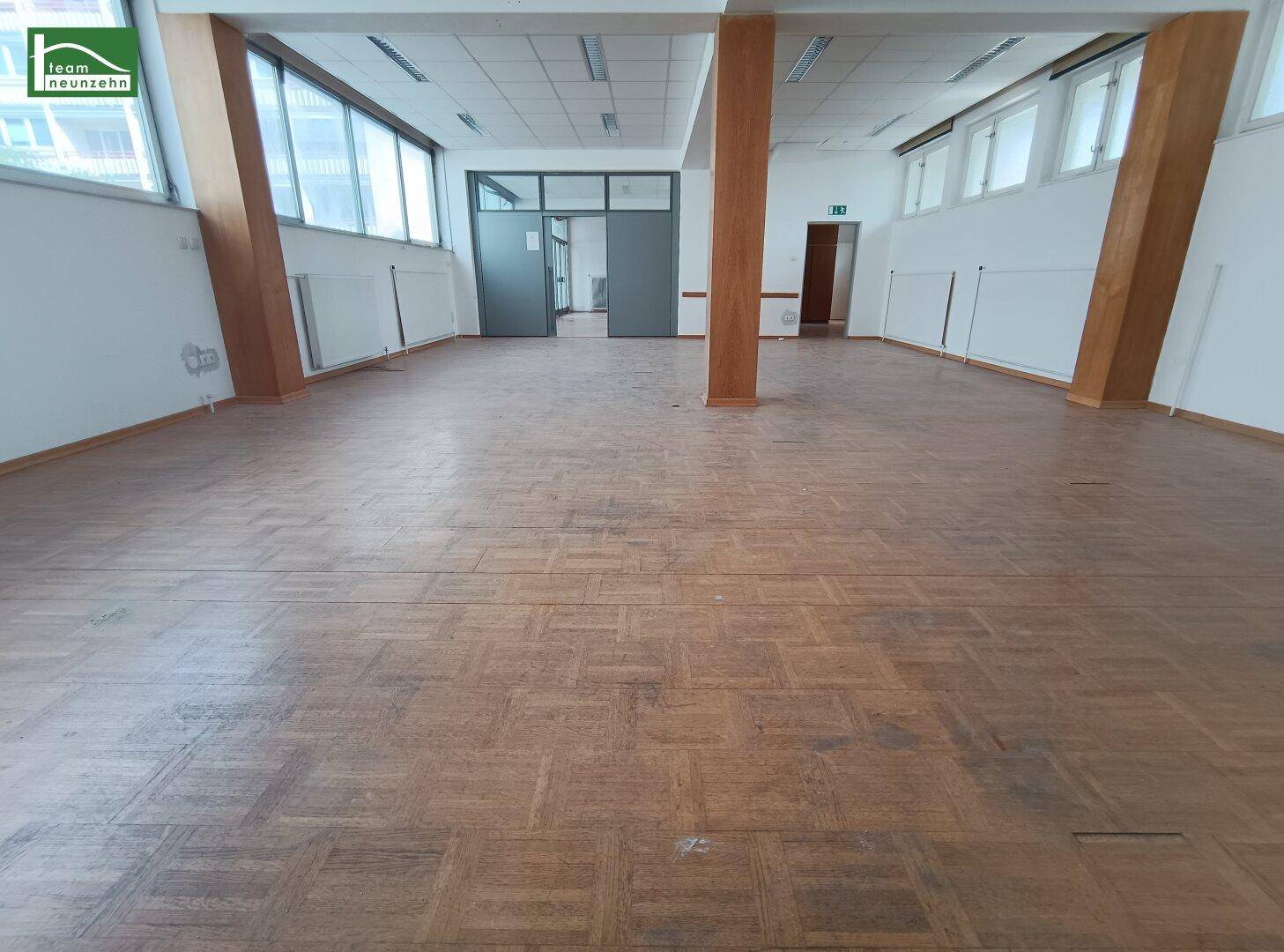 Geräumiger Bürobereich mit Parkettboden, großen Fenstern und Zugang durch Glastüren.