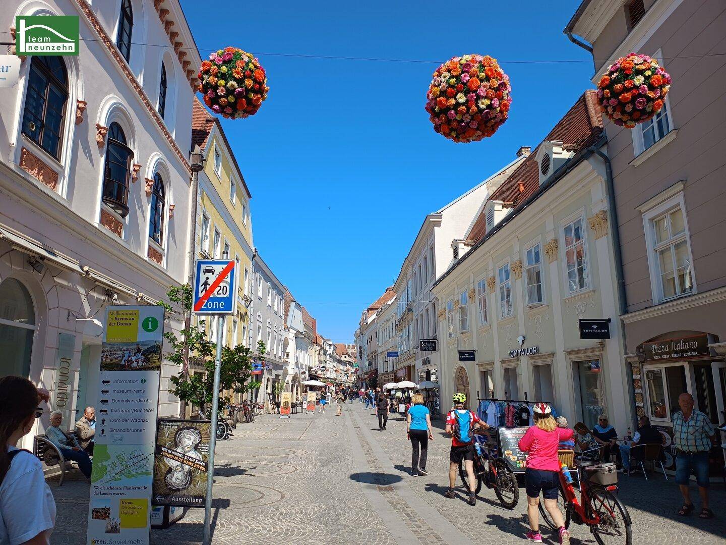 Belebte Einkaufsstraße in einer historischen Stadt mit dekorativen Blumenarrangements.
