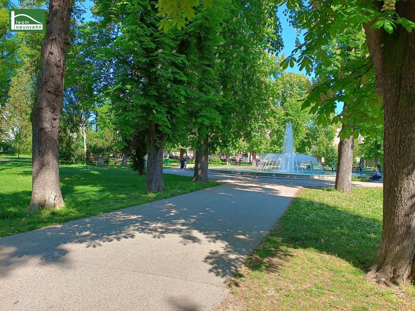 Grüner Park mit Bäumen, einem gepflasterten Weg und einem Springbrunnen im Hintergrund.