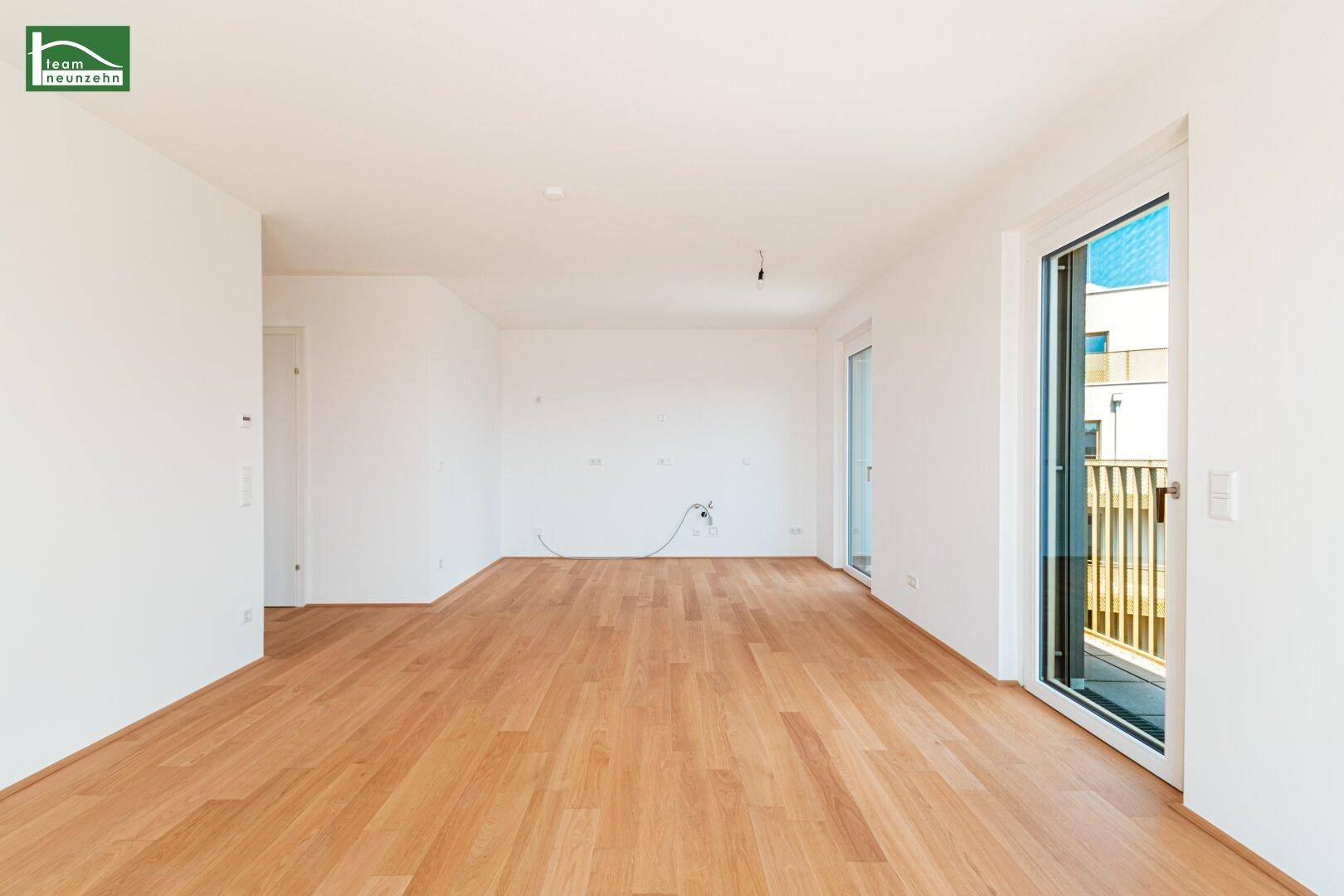Helles, leeres Wohnzimmer mit Parkettboden und Zugang zu einem Balkon.
